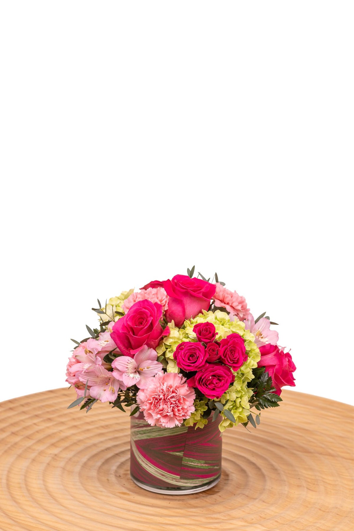 Tourmaline features hot pink roses, pink carnations, alstroemeria, and green hydrangea arranged in a glass cylinder vase wrapped with decorative leaves.