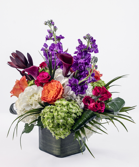 Tropical-inspired floral arrangement with purple calla lilies, hydrangea, roses, and dark purple stock in a clear cube vase