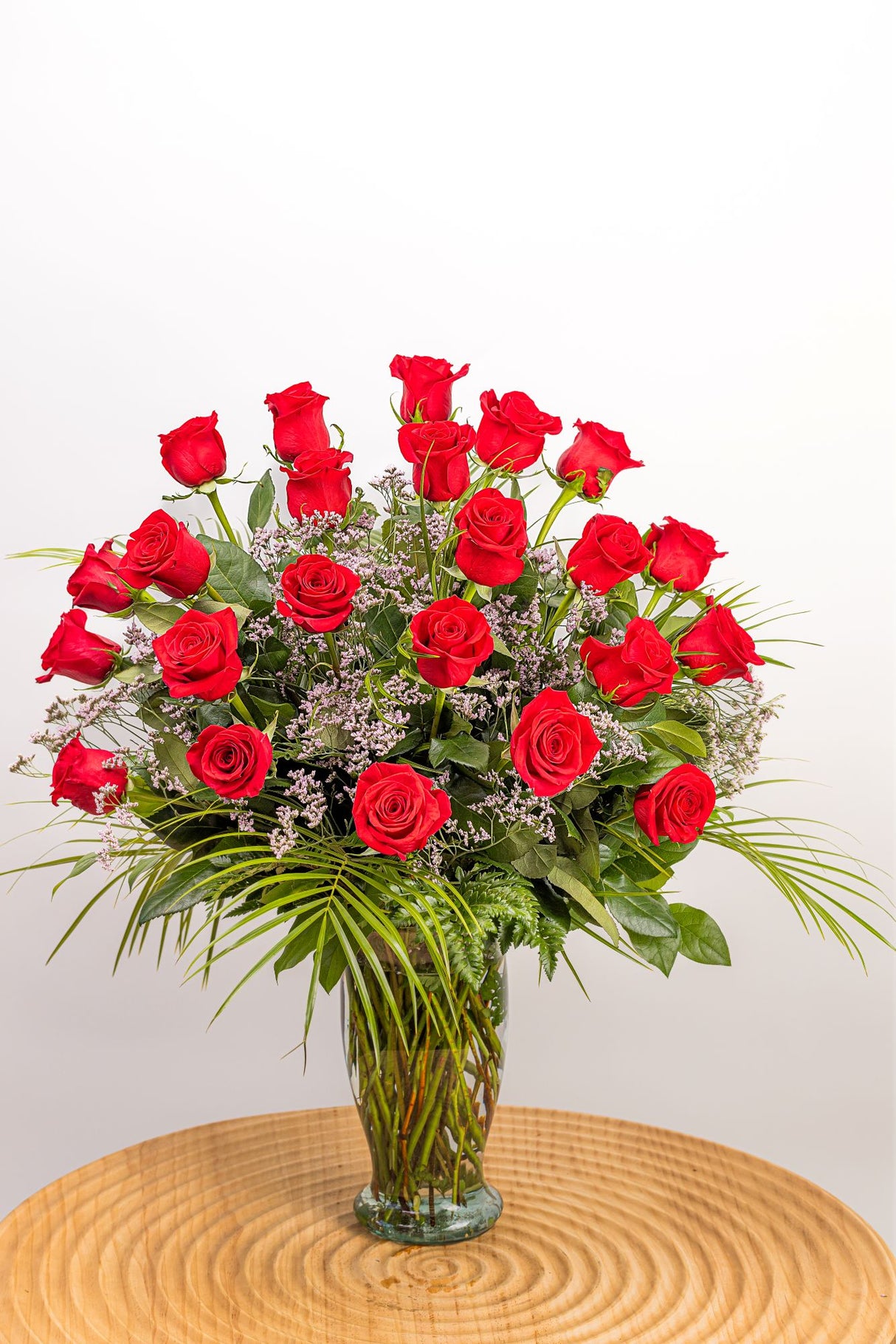 Two Dozen Premium Red Roses features twenty-four long-stem red roses accented with pink limonium, leatherleaf, salal tips, and robellini greenery, arranged in a ribbed glass vase.