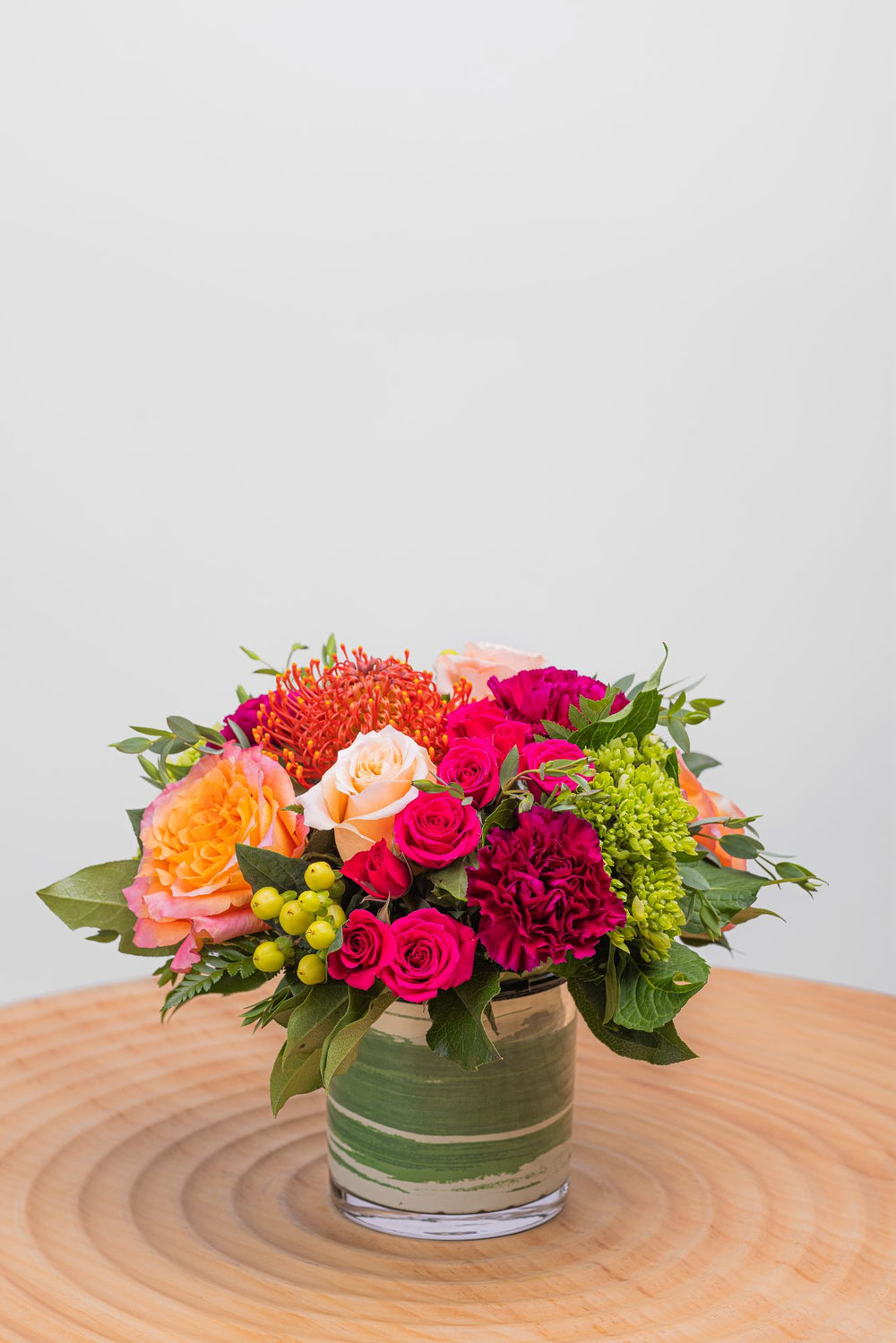 Velvet Petals features soft peach and Free Spirit roses, hot pink spray roses, purple carnations, orange pincushion proteas, mini green hydrangeas, hypericum berries, eucalyptus parvifolia and more greens in a cylinder glass vase with a ribbon leaf.