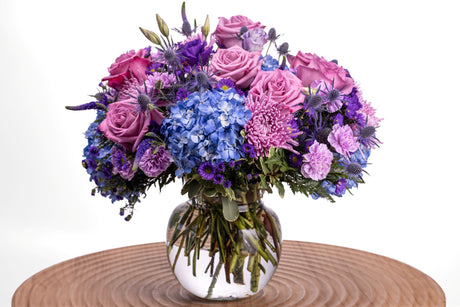 Violetta features lavender roses and carnations, purple lisianthus, veronica, asters, blue hydrangea, and thistle, arranged with greenery in a clear spittoon vase.