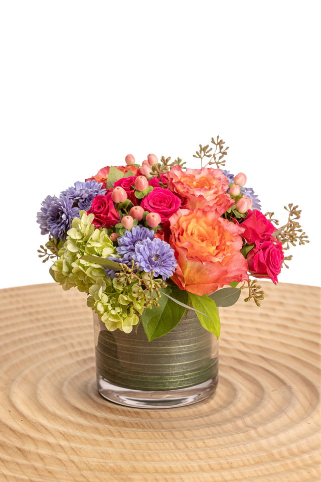Viva features Free Spirit roses, hot pink spray roses, green hydrangea, peach hypericum berries, blue cushion poms, seeded eucalyptus and salal in a glass cylinder vase.