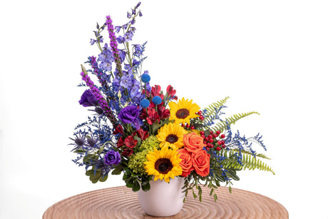 Vivid Blooms features sunflowers, orange roses, purple lisianthus, red alstroemeria, tinted craspedia, blue delphinium, and green hydrangea arranged in a white ceramic vase.